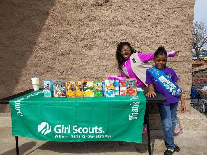 Girl Scouts smiling beside a table displaying boxes of cookies